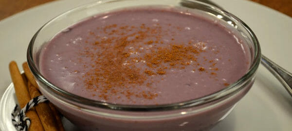 A clear bowl filled with blueberry soup on a white plate, topped with cinnamon, with a spoon and cinnamon sticks.