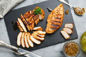 Sliced broiled chicken breast on a gray slate cutting board, with flatware, oil, and a bowl of spices.