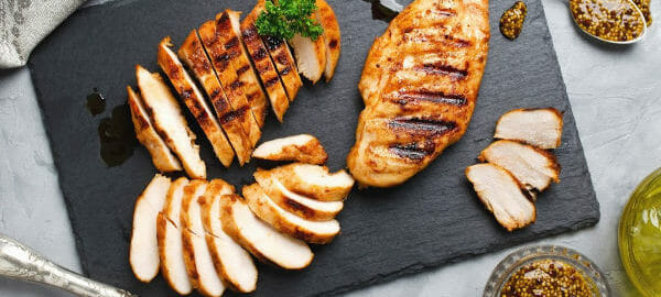 Sliced broiled chicken breast on a gray slate cutting board, with flatware, oil, and a bowl of spices.