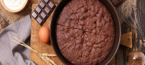 A round cake pan filled with a sliced Chocolate Mayonnaise Cake, with eggs, a whisk, and chocolate bars nearby.