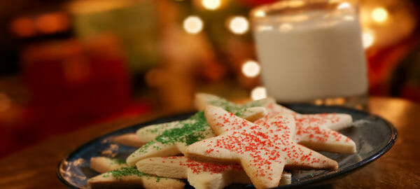 A black bowl filled with star-shaped cutout Cream Cheese Sugar Cookies, with a glass of milk at Christmastime.
