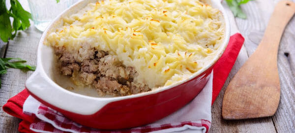 A red and white casserole dish on red and white towels, filled with Hamburger Pie that is topped with mashed potatoes.