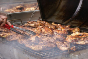Several pieces of Jerk Chicken on a hot grill and in a metal pan, with a person turning pieces with tongs.