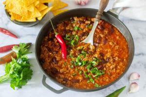 A gray metal skillet filled with Taco Chili, topped with a fresh chili pepper and shredded parsley, with corn chips nearby.