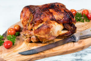 A whole browned Teriyaki Rotisserie Chicken on a wooden cutting board, with cherry tomatoes, parsley, and a knife.