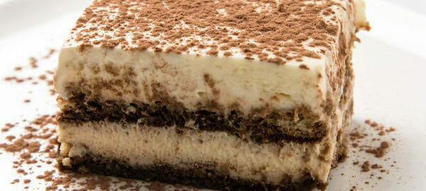 Close-up of a serving of layered Tiramisu on a white dessert plate, sprinkled with cocoa.