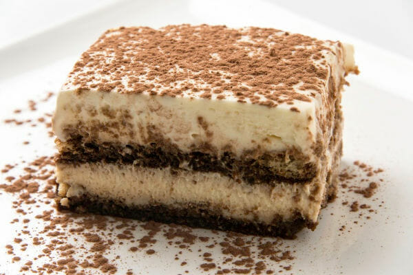 Close-up of a serving of layered Tiramisu on a white dessert plate, sprinkled with cocoa.
