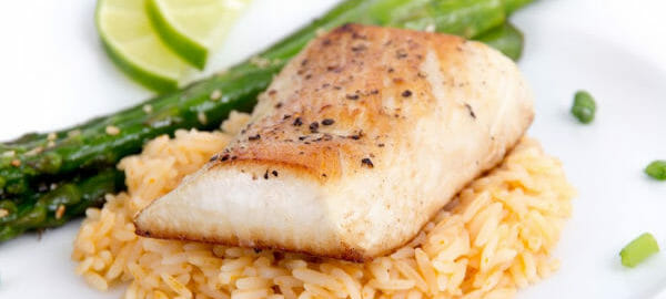 Close-up of a white plate with a serving of grilled tilapia on a bed of brown rice, with asparagus spears and lime slices.