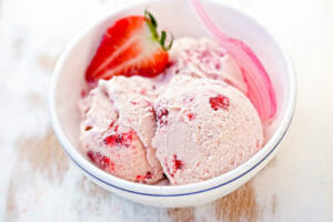 A white bowl filled with homemade strawberry ice cream, with a pink spoon and a fresh strawberry garnish.