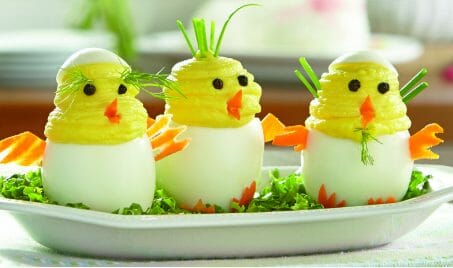 Three Deviled Eggs in the shape of chicks popping out of a white egg, with carrot wings and beaks, and black olive eyes.