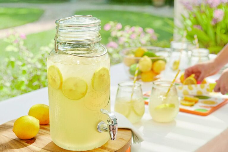 A glass drink dispenser filled with lemonade on a picnic table next to two glasses and citrus fruits